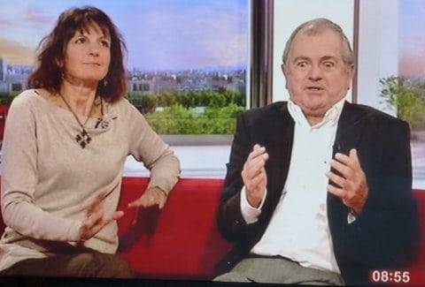 Prof and Helen Dewdney Woman brown shoulder length hair light brown jumper looking up to ceiling hand outs stretched, in agreement pose sitting on red sofa next to man in 50s white shirt blue jacket hand out in front foot apart. looking down in horror