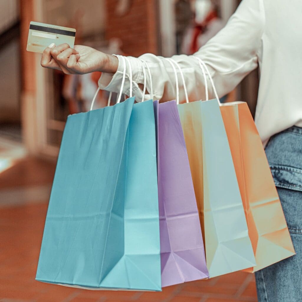 Arm in white jumper holding out credit card with 4 paper bags in light blue, light purple, aqua and orange with white handles over arm just see part of person's jeans