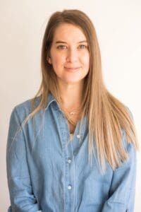 Woman in 40s long straight light brown hair smiling. Denim light blue shirt top two buttons undone