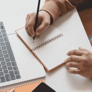 hand writing in notepad in front of laptop