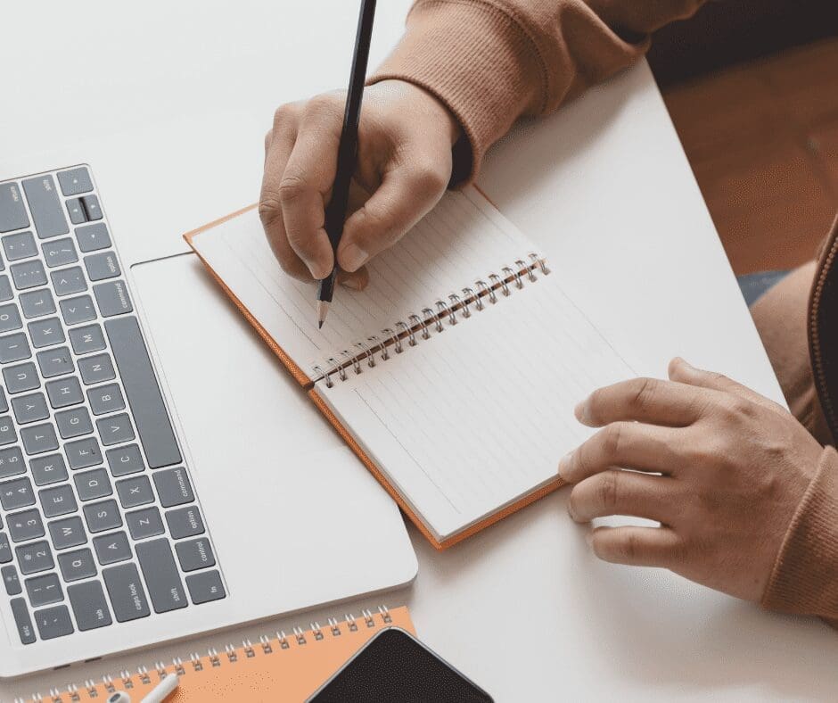 hand writing in notepad in front of laptop