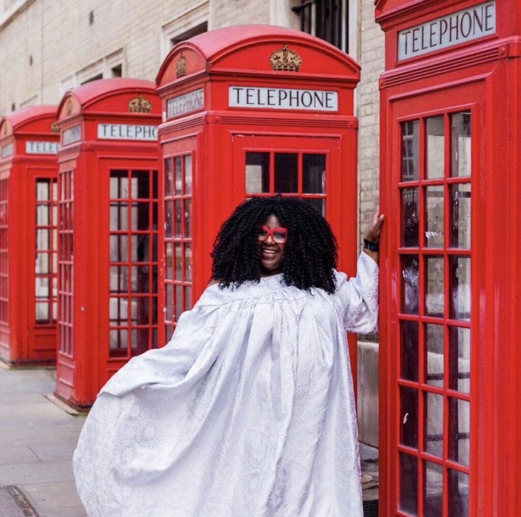 Black woman big long white dress between two of 4 old fashioned telephone boxes