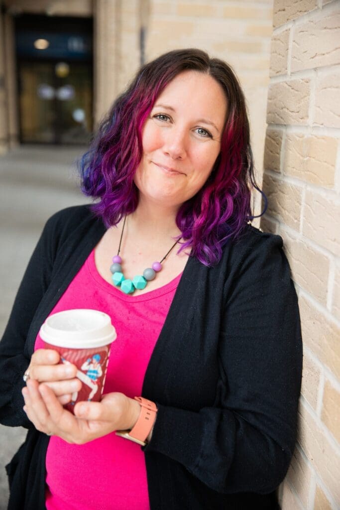 Woman leaning against wall dyed purple and blue should length hair. Pink top black jacket Holding take away coffee