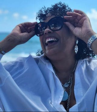 Black woman ands on sides of sunglasses laughing upwards thin white Summer top