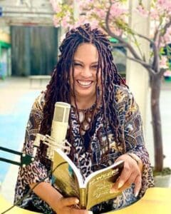 Black woman long dreadlocks reading book in front of microphone looking at camera