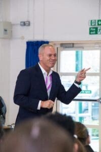 White man blue suit white shirt smiling pointing toward a member of the audience to the side and smiling towards the front