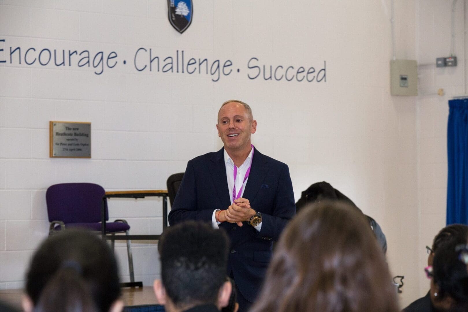 White man blue suit white shirt smiling at audience together hands together challenge and succeed and Heathcote on wall behind him