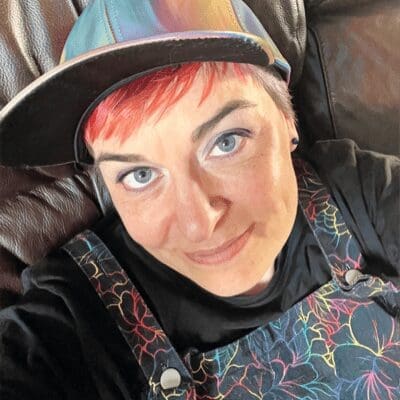 Woman's face looking up to camera, baseball cap on head red fringe hair black top colourful top of dungarees