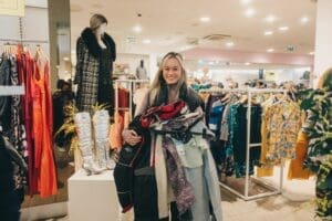Blond haired woman holding clothes on hangers in front of rail clothes and mannequin black coat and scarf with long white boots