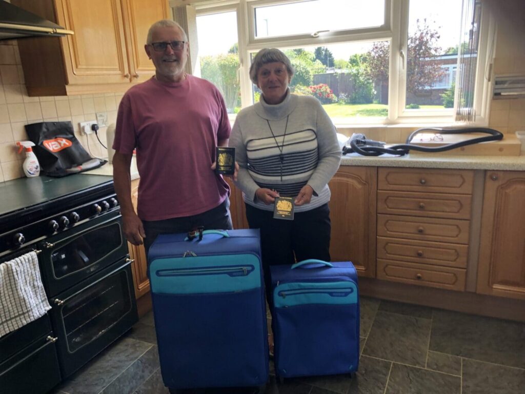 White couple in 60s standing up in kitchen behind two suitcases Man greay hair and beard sn light burgundy t shirt shorter woman in grey jumper