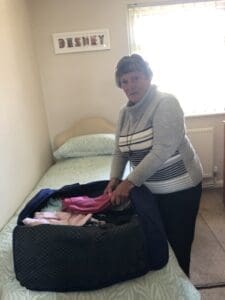 White woman in 60s short brown hair grey jumper packing suitcase on bed