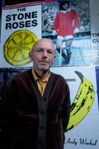 bald headed man in orange shirt black jacket standing in front of Stone Roses and footballer posters
