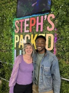 Steph Packed Lunch sign in background white woman in purple top next to black man in green top and denim jacket