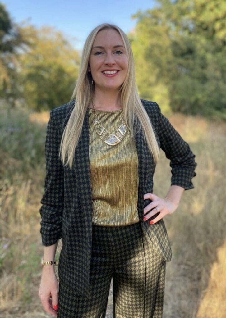 white woman long blond hair hand on hip. Standing in field long black and gold checked jacket and trousers gold top necklace and bangle