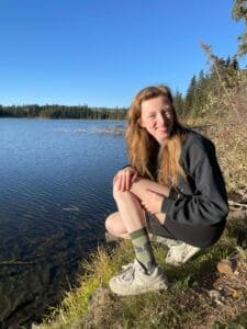 White woman crouching down in front of lake. Long light brown hair, smiling coat shorts and trainers with animal socks on