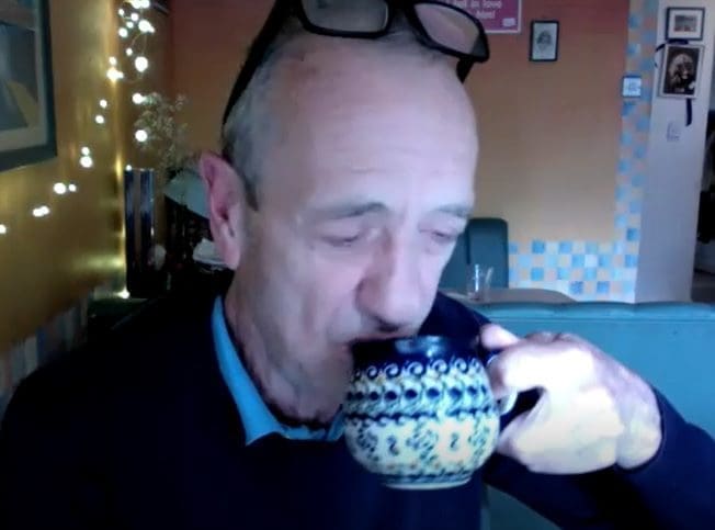 White man with alittle grey hair black rimmed glasses on head eyes shut drinking from mug, navy jumper light blue shirt collar underneath