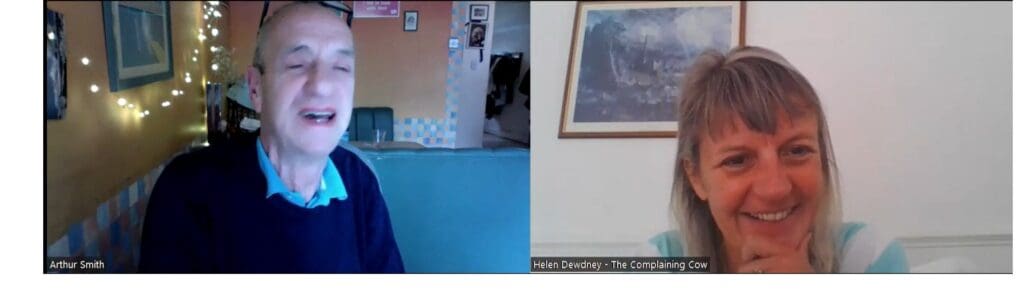 Zoom screenshot white man late 60's nearly bald black rimmed glasses on head navy blue jumper and light blue collared shirt. White woman briwn/grey hair mid 50s white and light green striped top sitting on sofa