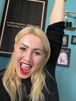 White woman in mid 40s, long straight blond hair. black top raised left arm open mouthed cheering blue wall with pictures with black frames
