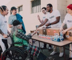 white man, black main in white t shirts with "volunteer" on them behind table with drinks on man in wheelchair long gey hair green checked top Asian man white man with him