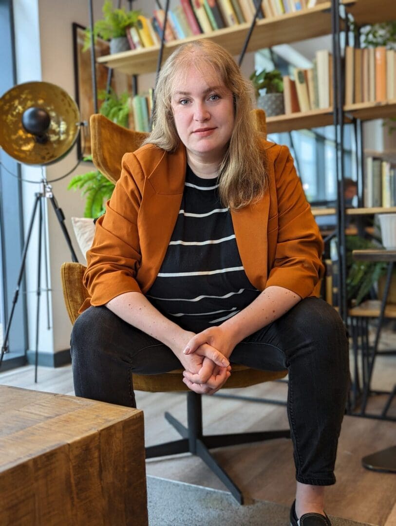 White woman blond hair sitting on a chair dark blue trousers horizontal dark blue with thin white blue stripes camel colour jacket
