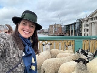 White woman outside long straight brown hair black hat blue scarf and grey jacket smiling at camera 5 sheep behind her