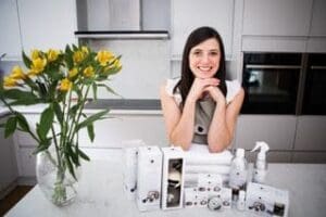 White woman long straight brown hair head on hand elbows on table smiling, wool products in front of her vase of yellow flowers to left facing background kitchen