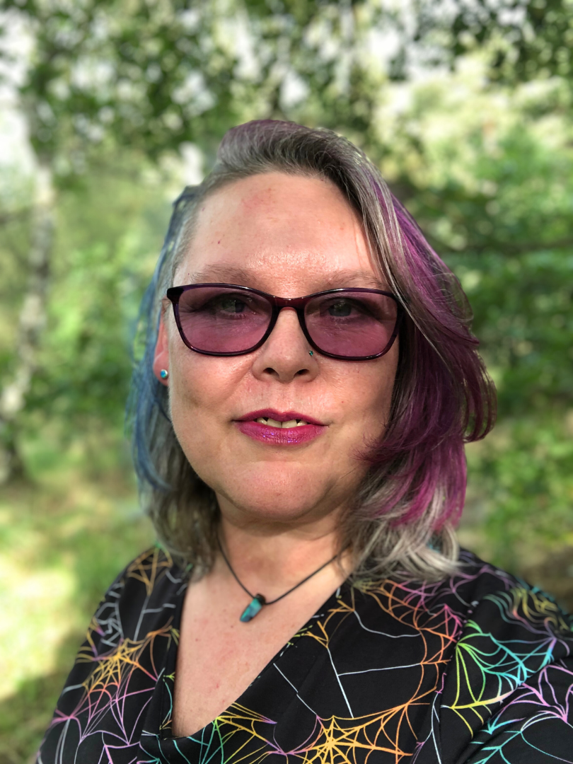 White woman looking at camera head shot, in front of a tree. Sunglasses dyed purple hair