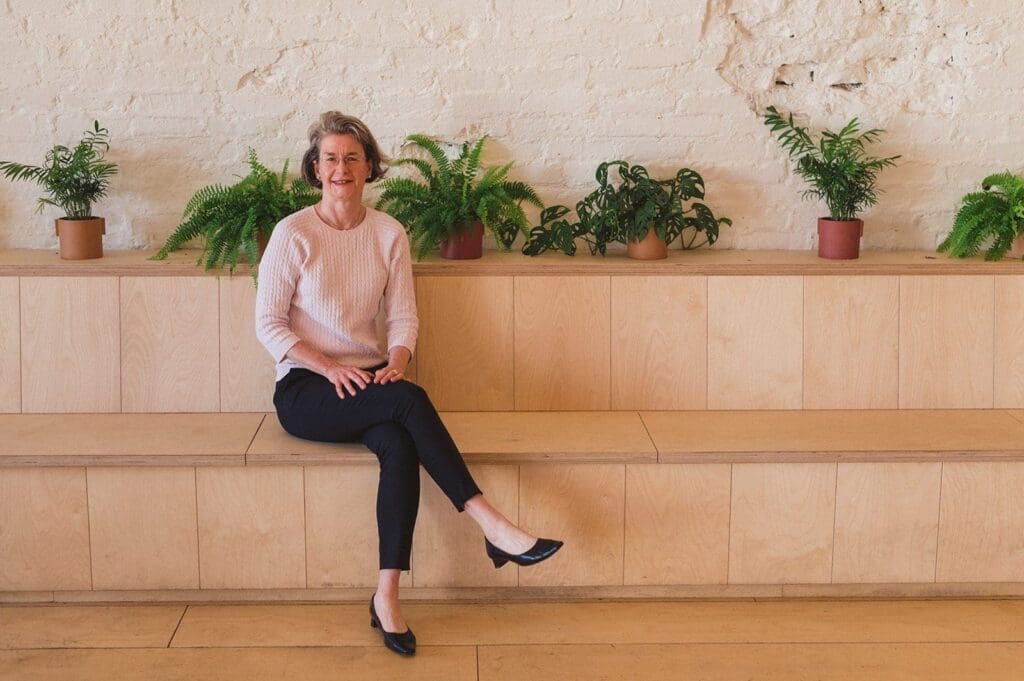 White woman sitting on step with greenery on the second one behind her. Short dark shoulder length hair, smiling, bilk long sleeve top cross leg black leggins low heeled dark shoes cross legged