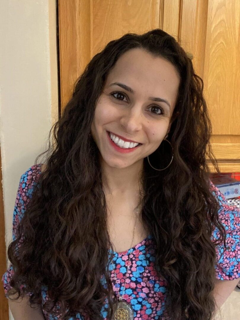 Head shot white woman long dark brown wavy hair, blue ad pink top. Smiling in front of kitchen cupboard