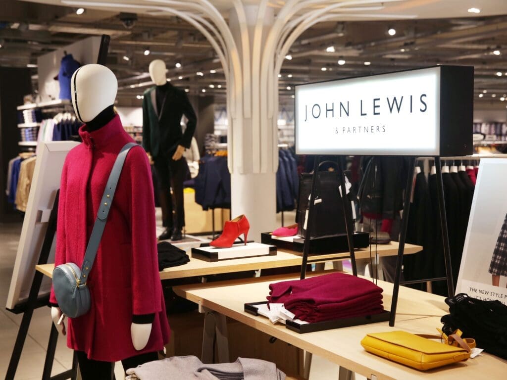 John Lewis sign in store, mannequin in front of sign and table with a red bag on it. Red winter coat on mannequin done up no buttons showing black handbag across body