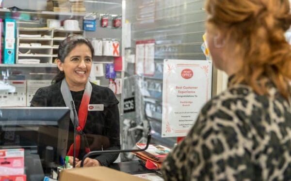 White woman dark hair tied back smiling black top name badge and lanyard serving white woman in leopard print top blond hair half side shot as being served through glass.