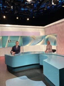 Channel 5 News studio Dan behind the desk Helen to side. Dan slim man in blue suit and tie smiling short brown hair, Helen long sleeve spotted cream and brown top smiling shoulder length wavt blond/grey hair