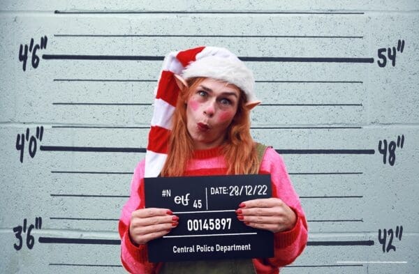 White woman against background with lines showing 4ft high,. Pulling a face pink and red striped top, long white and red stripe hat. Holding a black board with 28/12/24 and police number