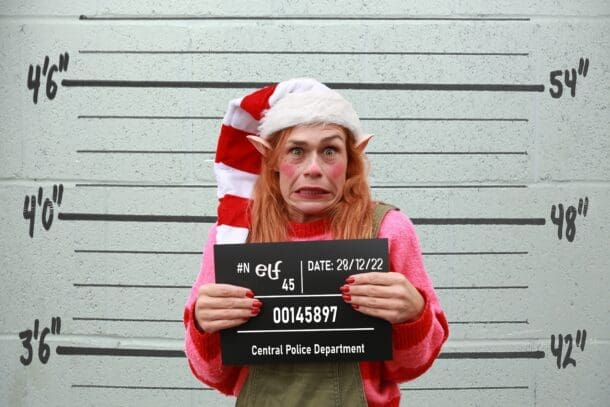 White woman against background with lines showing 4ft high,. Pulling a face pink and red striped top, long white and red stripe hat. Holding a black board with 28/12/24 and police number