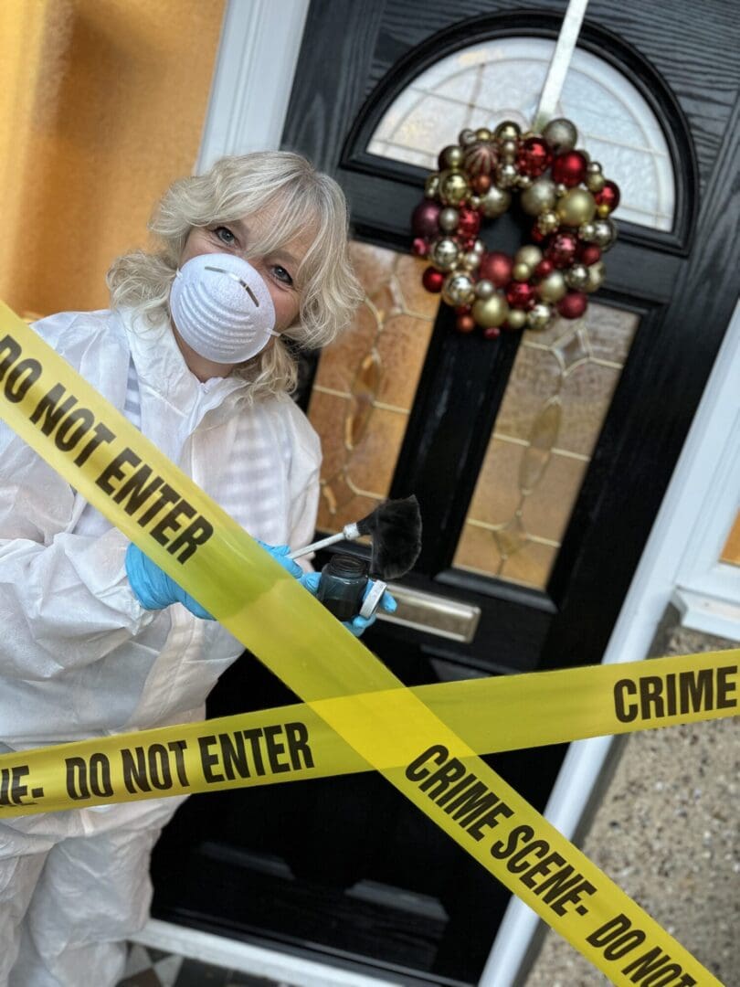 Woman in early 50s short wavy blond white hair with mask standing in front of front door with red and gold bauble balls wreath Do not enter yellow tape crossed in front.