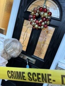 Woman in early 50s short wavy blond white hair with mask standing in front of front door with red and gold bauble balls wreath swabbing letterbox, Do not enter yellow tape crossed in front.