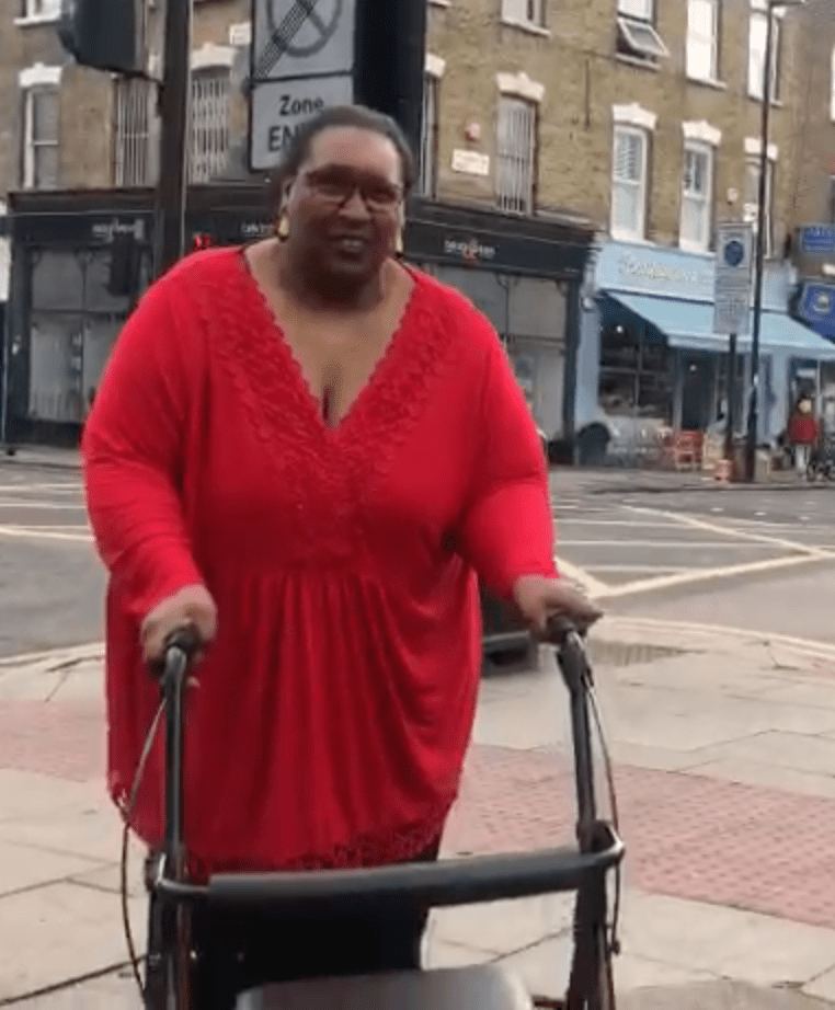 Black woman with glasses big loose red top, smiling holding walking frame