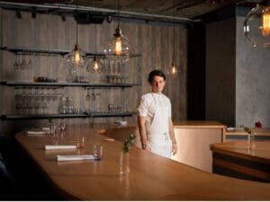 White man black hair in kitchen whites standing behind long wooden table with glasses on it curving round him part of wooden table to left of him, glasses on shelves behind him