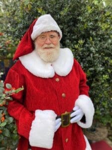 Santa red coat white cuffs bit white fluffy collar hat white band red length behind. White beard glasses hands with white gloves on black belt round his middle in front of a bush