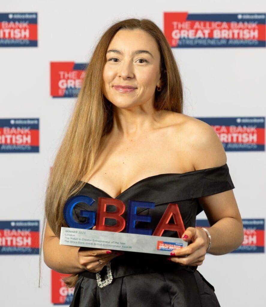 White woman long straight light brown hair, smiling towards camera. Holding award. Wearing off shoulder black shiny dress