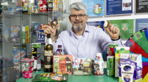 White man with grey hair and geard and glasses. Smiling, white shirt behind various products
