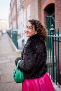 White woman walking along street looking back at camera smiling. Shoulder length brown curly hair fur jacket green handbag on left arm