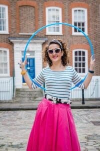 White woman in lates 20s holding up a blue hoola hoop behind her. Curly shoulder length brown hair green and white stripe see the top of pink skirt. Standing in front of terrace houses