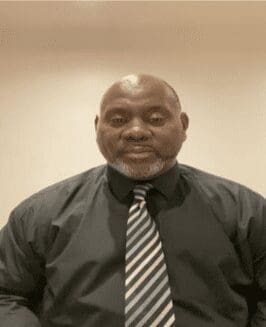 Black man bald short grey bears brown shirt and brown and cream striped tie, neutral face