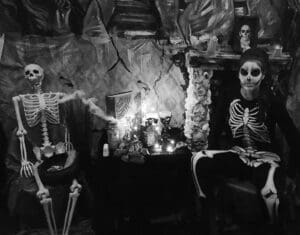 Black and white picture. Skelton to the left someone dressed up and made up as skeleton to right in front of cobwebs and table with lights, stands with creepy faces