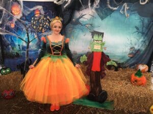 Woman in late 20s smiling hair tied back wearing dress with long sleeved dark turquoise long sleeve yellow lacing up the front big puf ball orange netting layered skirt. Tree in background hay on floor with pumpkins, blue background with cobwebs a fun Frankenstein figure/statue behind green face red t shirt and black trousers and jacket green hands outstretched