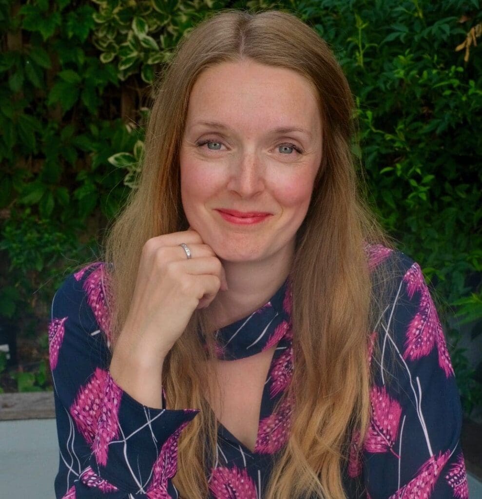 Head shot woman in pink and blue flowery top head resting on right hand in fist. Long straight light brown hair white woman