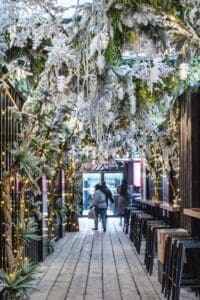 Long bar with stools to the right white/grey square tiled floor double window doors at the end figure near the doors. Trees with white blossom making arch