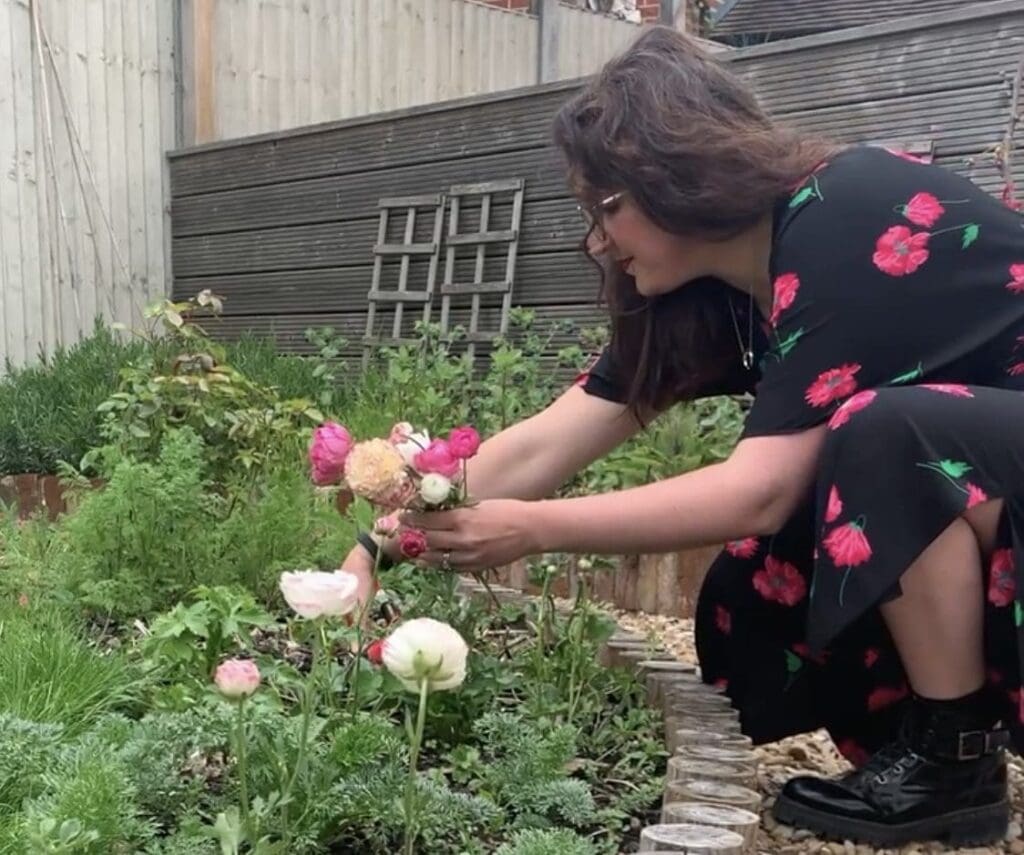 Woman in 30s blank and pink flowers dress, long straight brown hair bending down to cut flowers from shrubery