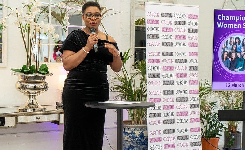 Black woman in late 30s long black dress short sleeves. Glasses short black hair standing at a round table. Talking into a microphone. Display Stand with logos on it to the side. Inbetween just behind her large white and blue plant pot with green grass plant. Window behind her.